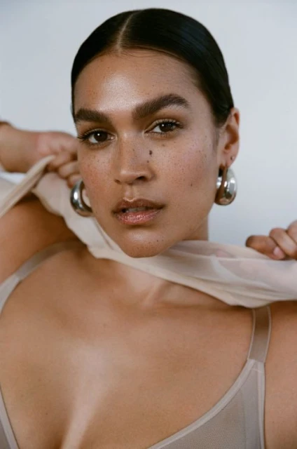 Close-up portrait of a woman with dark hair, fair skin, hoop earrings, and a beige top, posing against a neutral background in soft lighting.