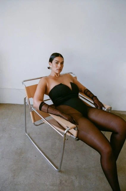 Woman in a black bodysuit and sheer tights poses on a sling chair in front of a white wall. She has black hair and tan skin.