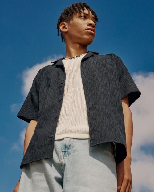 Young man poses in a dark blue patterned shirt and light blue denim shorts against a bright blue sky with scattered clouds.