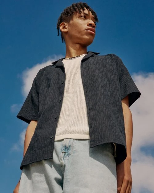 Young man poses in a dark blue patterned shirt and light blue denim shorts against a bright blue sky with scattered clouds.