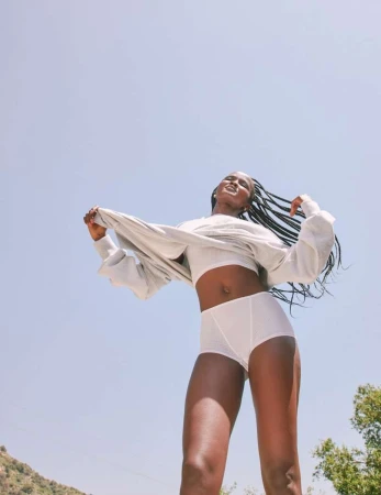 Black woman with long braids wearing white underwear and a matching top under a light sweater outdoors