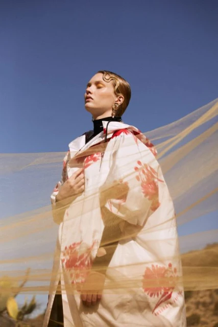 Young adult woman in floral dress with golden veil stands in desert against blue sky. Fashion photography with fine art and editorial style.
