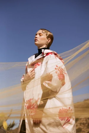 Young adult woman in floral dress with golden veil stands in desert against blue sky. Fashion photography with fine art and editorial style.