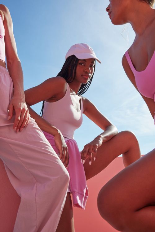Three women in pink athletic wear are outdoors in bright sunlight against a blue sky, showcasing fashion and fitness lifestyle.