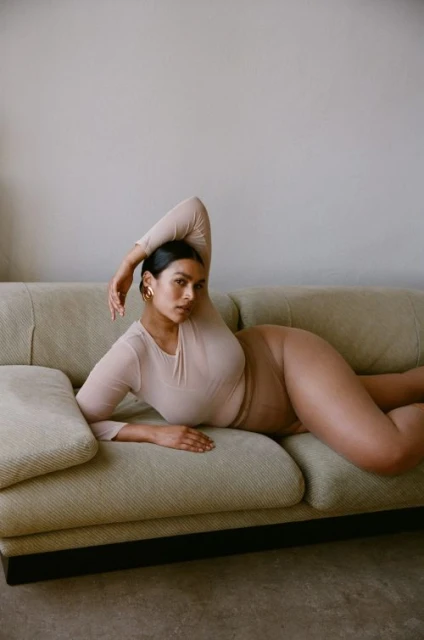 Adult woman in a neutral-toned bodysuit lounging on a couch in a relaxed pose, photographed indoors against a neutral wall.