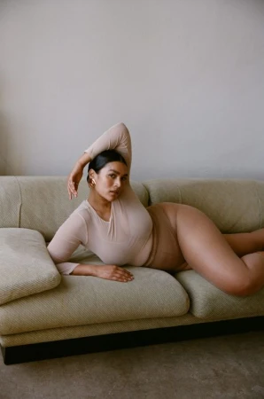 Adult woman in a neutral-toned bodysuit lounging on a couch in a relaxed pose, photographed indoors against a neutral wall.