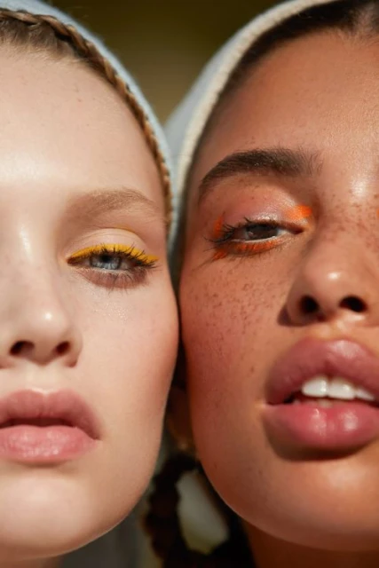 Close-up portrait of two women with contrasting skin tones, wearing headscarves and vibrant makeup, set against a soft, blurred background.