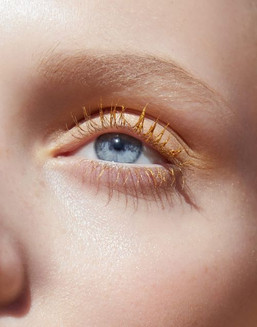 Close-up of a light-skinned adult female with bright blue eye and golden mascara, fair eyebrows and skin, illuminated by natural light.