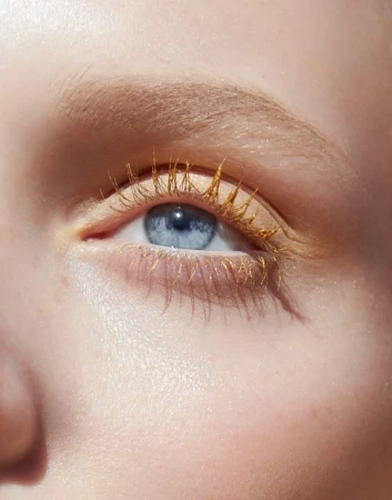 Close-up of a light-skinned adult female with bright blue eye and golden mascara, fair eyebrows and skin, illuminated by natural light.