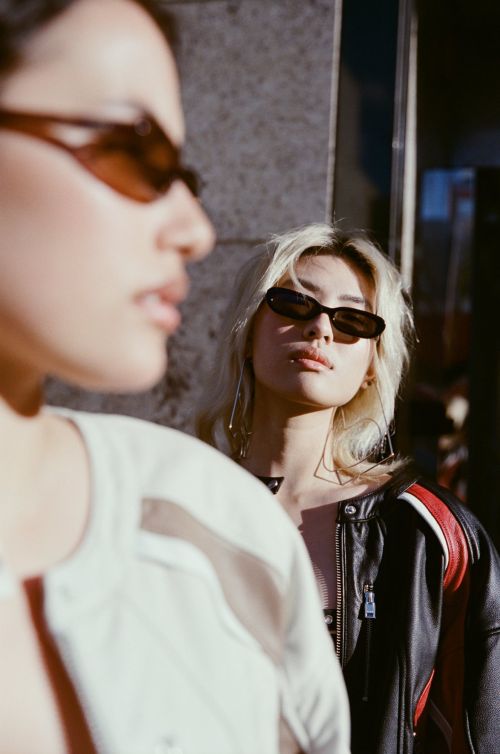 Two Asian women with light hair and sunglasses in leather jackets pose against a concrete background with soft, diffused sunlight.