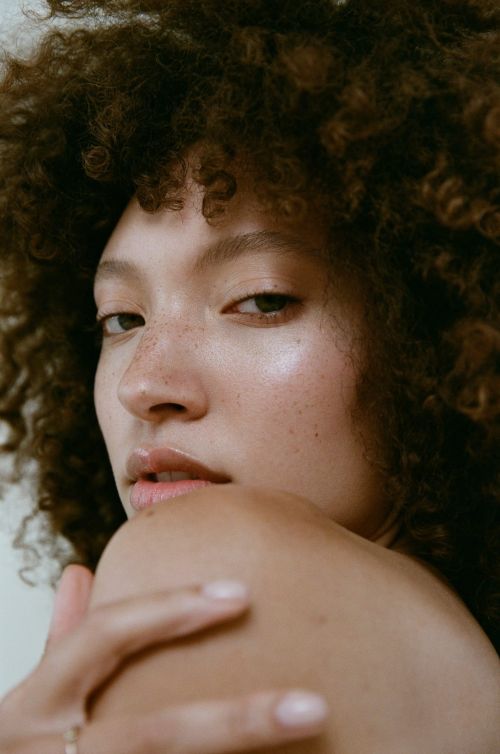 Close-up portrait of a young adult woman with curly hair and a serene expression in a studio setting with soft, diffused lighting.