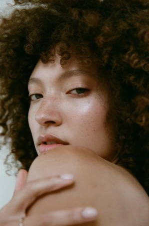 Close-up portrait of a woman with curly brown hair, freckles, and natural makeup looking over her bare shoulder.
