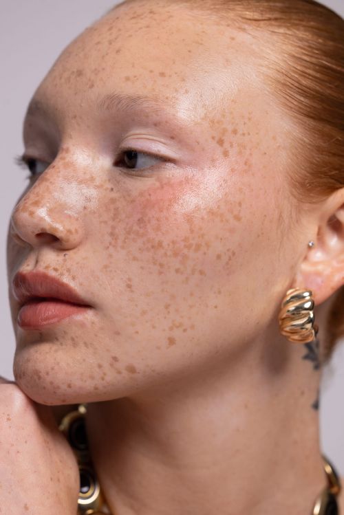 Close-up portrait of a young adult female with freckles and golden hoop earrings in a studio setting with neutral background.