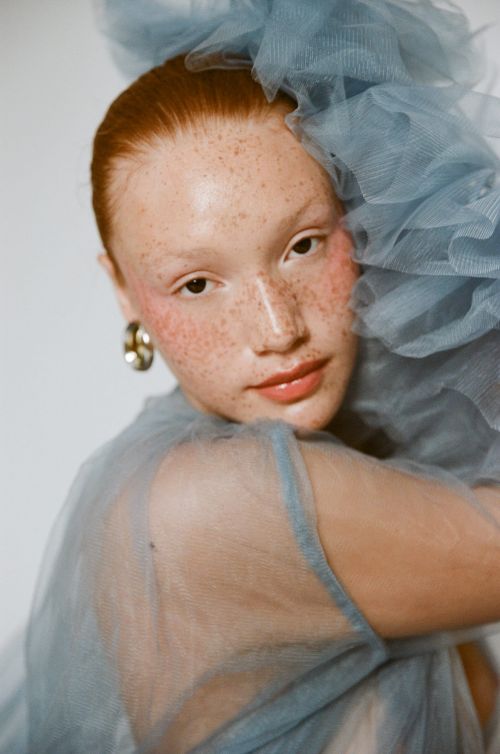 Portrait of a red-haired woman with freckles wearing a blue tulle dress, looking at the camera against a white background.