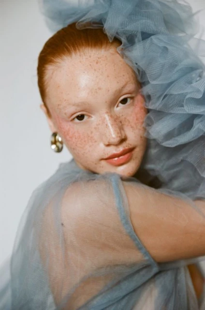 Portrait of a red-haired woman with freckles wearing a blue tulle dress, looking at the camera against a white background.