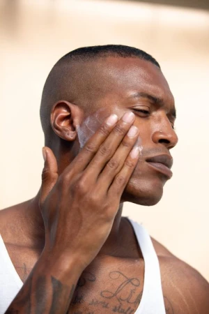 Close-up of an African American man applying sunscreen or moisturizer to his face, wearing a white tank top against a neutral background.