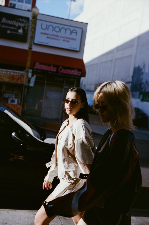 Two adult women with sunglasses walk on an urban street near buildings and a parked car, bathed in bright daylight.