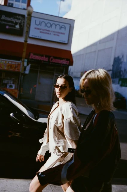 Two adult women with sunglasses walk on an urban street near buildings and a parked car, bathed in bright daylight.