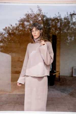 Woman wearing a matching taupe turtleneck sweater and long skirt posing outdoors near trees and buildings