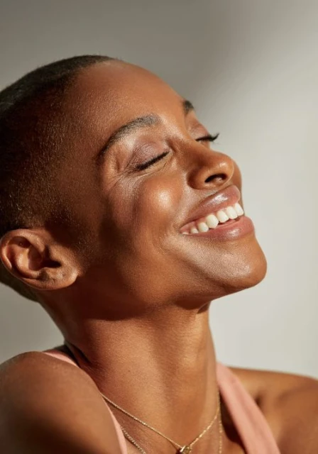 Portrait of a smiling Black woman with a buzzcut and a golden necklace, eyes closed, head tilted, basking in soft, bright, natural light.