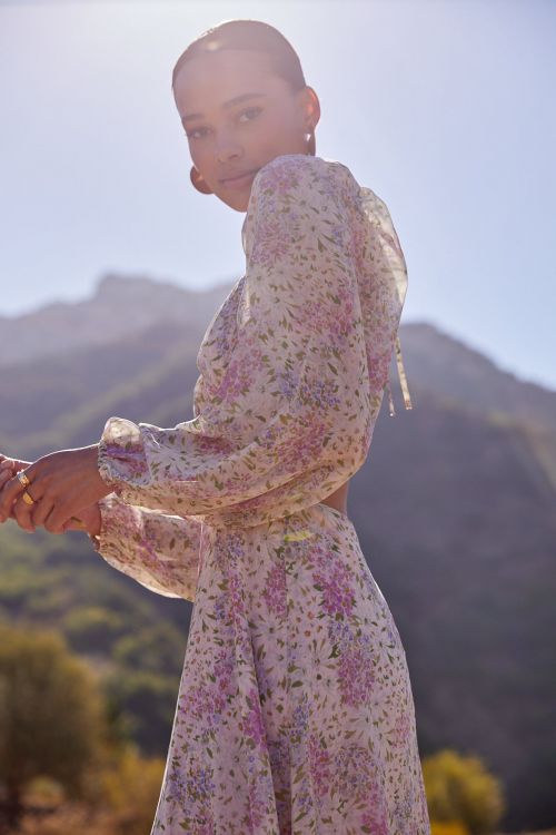A woman poses in a long, floral print dress in an outdoor setting with mountains and blue skies in the background on a sunny day.