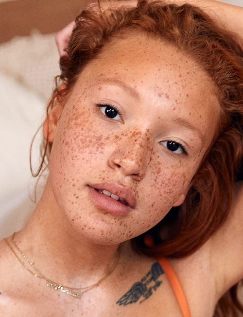 Close-up portrait of a freckled redhead woman with gold jewelry and a shoulder tattoo, set against a neutral indoor backdrop.