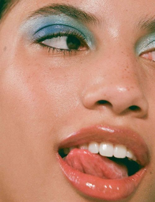 Close-up of a woman's face with blue eyeshadow and glossy lips, showcasing beauty makeup and a playful expression with tongue.