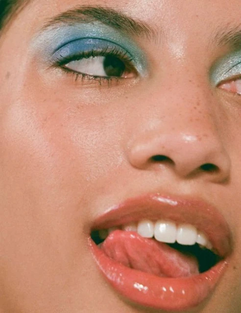 Close-up of a woman's face with blue eyeshadow and glossy lips, showcasing beauty makeup and a playful expression with tongue.