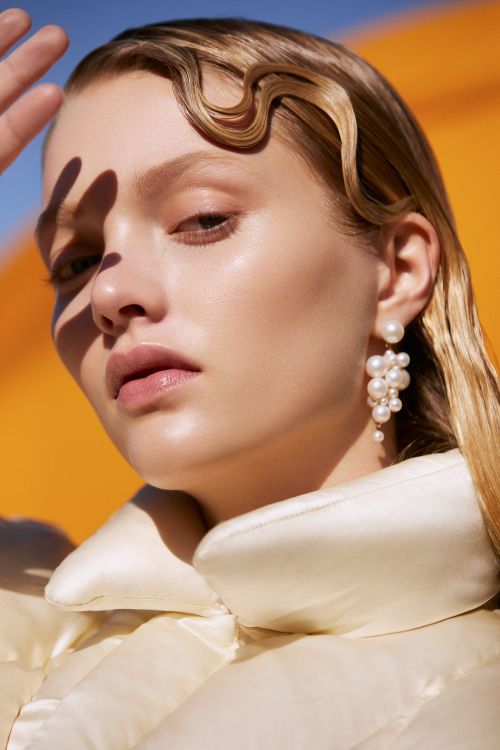Close-up of a light-skinned young woman with blonde wavy hair, pearl earrings, and a cream-colored coat, against an orange background.
