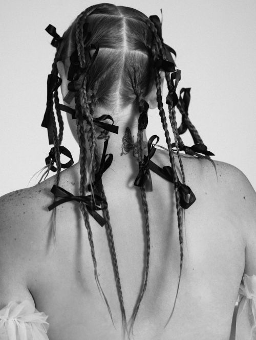 Back view of a person with blonde braided hair adorned with black ribbons and a butterfly tattoo on their neck, against a white background.