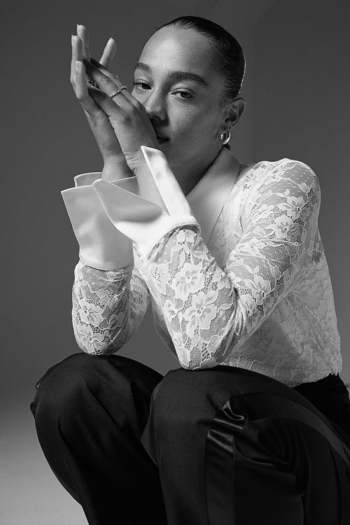 Portrait of a woman with hands near her face, wearing a lace long-sleeve shirt and dark pants, captured in a black and white studio setting.