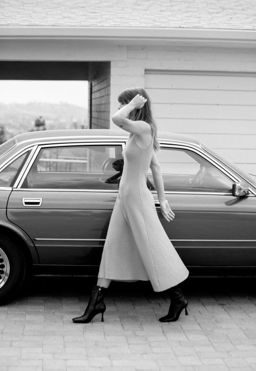 A woman in a dress walks past a vintage car in a monochrome outdoor shot, showcasing fashion and automotive elements.