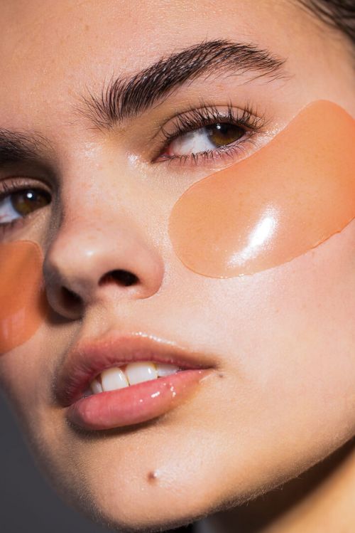 Close-up portrait of a woman with peach-colored under-eye mask patches, glossy lips, and neutral makeup against a neutral background.