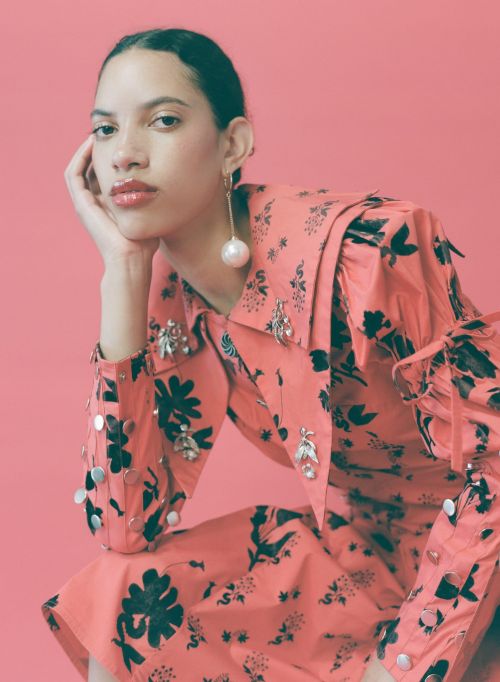 Young woman with dark curly hair poses in a coral-colored floral dress with pearl earrings against a solid pink background.