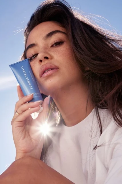 Brunette woman with fair skin holds a blue Summer Fridays skincare tube in a close-up shot against a light blue sky backdrop.