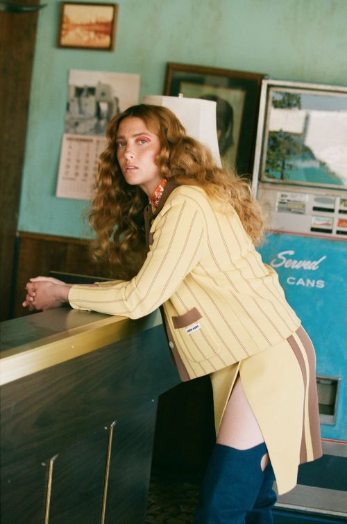 Fashion shot of a woman with auburn hair wearing a yellow-and-brown striped skirt and jacket, standing indoors next to a machine.