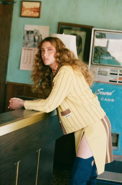 Fashion shot of a woman with auburn hair wearing a yellow-and-brown striped skirt and jacket, standing indoors next to a machine.