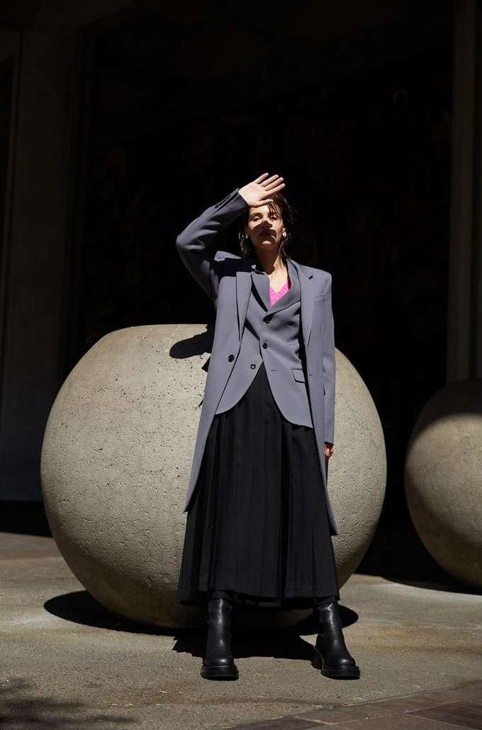 A woman in a grey blazer and black skirt stands in front of a dark background with large round planters in natural light.