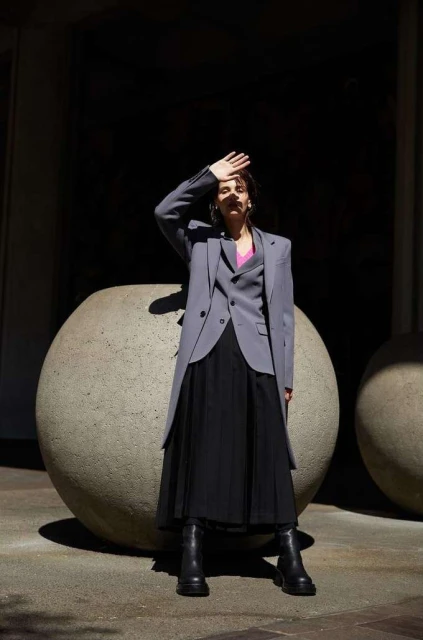 A woman in a grey blazer and black skirt stands in front of a dark background with large round planters in natural light.