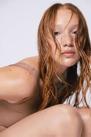 Close-up portrait of a woman with red hair and freckles wearing a nude tank top against a white background