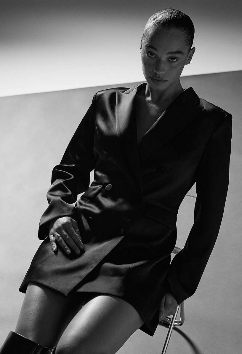 Studio portrait of an adult female wearing a black blazer dress and boots, seated on a chair with a neutral background.