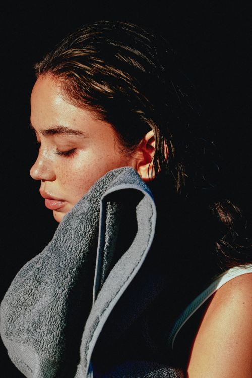 Close-up profile of a woman with wet hair drying her face with a grey towel against a dark background
