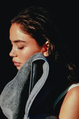 Close-up profile of a woman with wet hair drying her face with a grey towel against a dark background