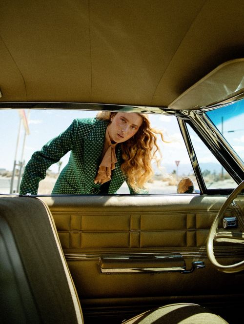 Woman in green jacket leans from inside vintage car; desert landscape and gas station visible through the front window.