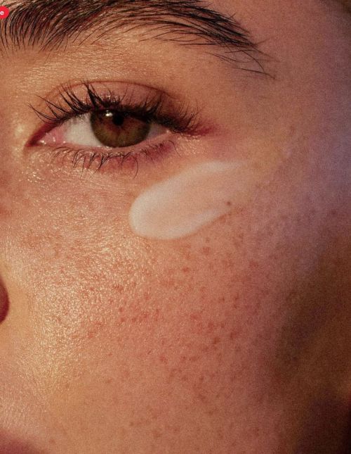Close-up shot of a person's face with light brown eyes and visible eyelashes, applying white cream or lotion on the cheek.