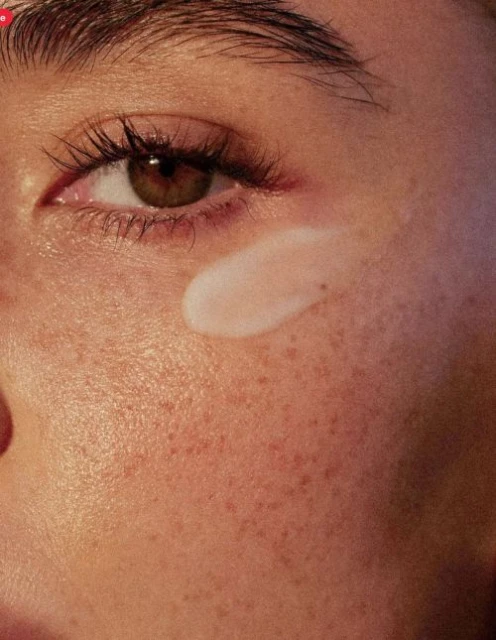 Close-up shot of a person's face with light brown eyes and visible eyelashes, applying white cream or lotion on the cheek.