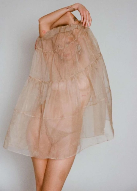 Woman in a sheer layered dress covering her head, posing indoors with minimalist white wall background, artistic fashion portrait.