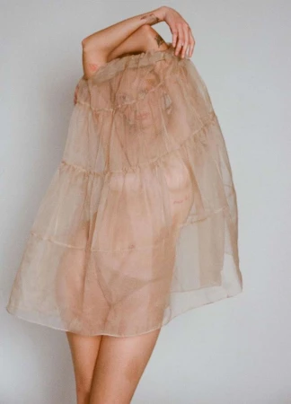Woman in a sheer layered dress covering her head, posing indoors with minimalist white wall background, artistic fashion portrait.