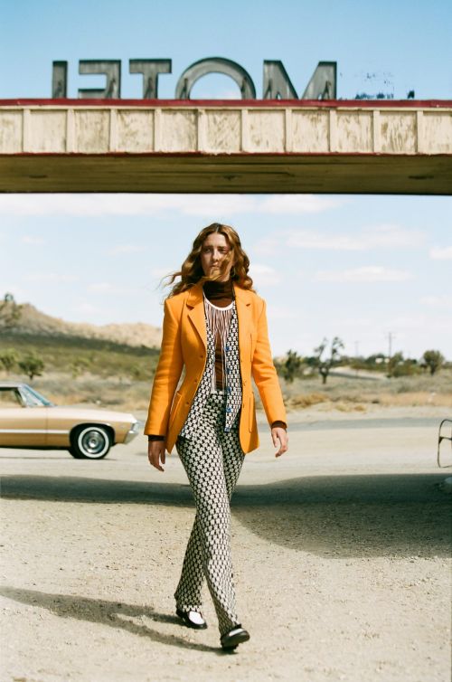 Model in orange blazer and patterned pants walks confidently on a desert road, with a vintage car and motel sign in the background.