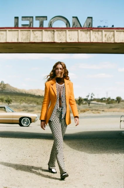 Model in orange blazer and patterned pants walks confidently on a desert road, with a vintage car and motel sign in the background.